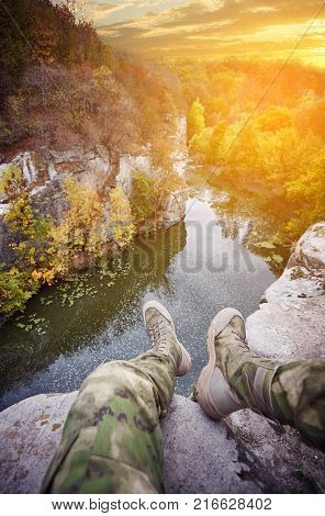 Traveler at a mountain river, legs over a cliff, outdoor clothing, deep stone canyon, ancient granite rocks, beautiful sunset, rocky river bank, cloudy sky, mountain landscape, forest on the riverbank
