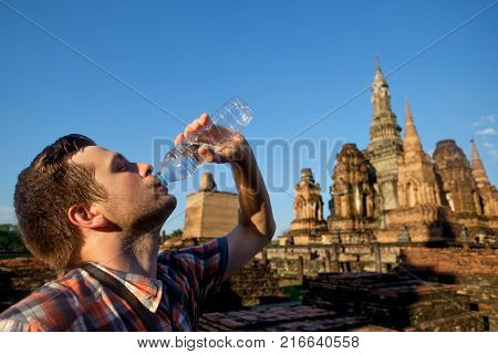 Young man thirsty is drinking bottled water in buddhism temple in Thailand on hot sunny weather