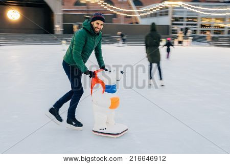 Outdoor Shot Of Glad Bearded Male Wears Green Jacket And Hat, Stands Near Skate Aid As Trying To Obt
