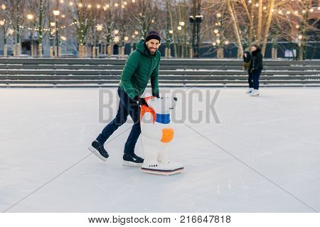 Outdoor Shot Of Unexperienced Male Skater Uses Skate Aid For Go Skating On Ice, Being In Good Mood,