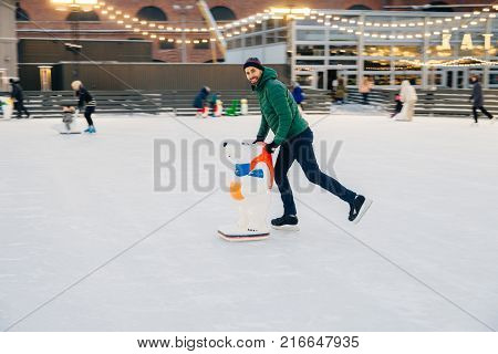 Outdoor Shot Of Unexperienced Male Skater Uses Skate Aid For Go Skating On Ice, Being In Good Mood,