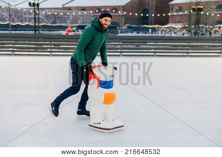 Outdoor Shot Of Unexperienced Male Skater Uses Skate Aid For Go Skating On Ice, Being In Good Mood,