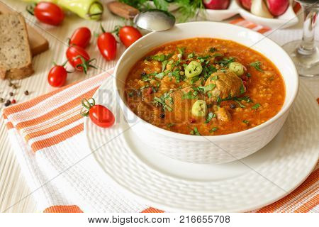 Traditional Georgian soup Kharcho with meat and rice in a white bowl next to bread, tomatoes, peppers, herbs on a white orange tablecloth. Horizontal. Close-up.