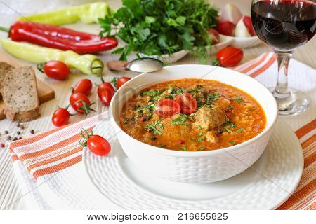 Traditional Georgian soup Kharcho with meat and rice in a white bowl next to bread, tomatoes, peppers, herbs, a glass of red wine on a white orange tablecloth. Horizontal. Close-up.