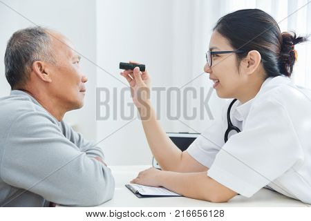 Young Asian Female Doctor wear glasses checking senior man patient eyes with flashlight in medical office.