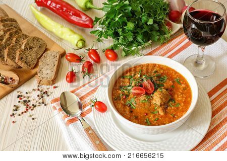 Traditional Georgian soup Kharcho with meat and rice in a white bowl next to bread, tomatoes, peppers, herbs, a glass of red wine on a white orange tablecloth. Horizontal. View from above.