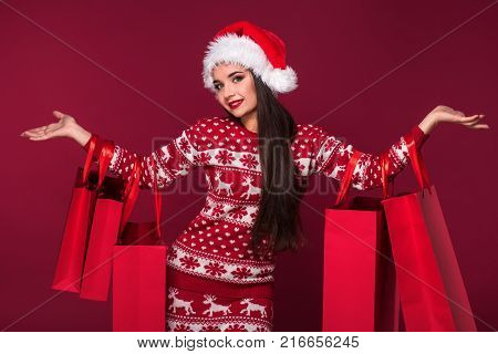 Young pretty woman or girl with long beautiful hair in Santa hat and New Year's dress holding red package on red studio background. Christmas sale. Emotional brunette. Copy space