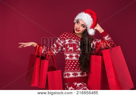 Young pretty woman or girl with long beautiful hair in Santa hat and New Year's dress holding red package on red studio background. Christmas sale. Emotional brunette. Copy space