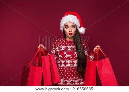 Young pretty woman or girl with long beautiful hair in Santa hat and New Year's dress holding red package on red studio background. Christmas sale. Emotional brunette. Copy space