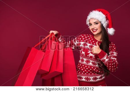Young pretty woman or girl with long beautiful hair in Santa hat and New Year's dress holding red package on red studio background. Christmas sale. Emotional brunette. Copy space