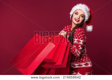 Young pretty woman or girl with long beautiful hair in Santa hat and New Year's dress holding red package on red studio background. Christmas sale. Emotional brunette. Copy space