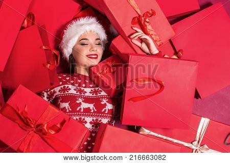 Funny young woman in a New Year's dress lying on a mountain of gifts. Christmas sale. Emotional brunette in Santa hat