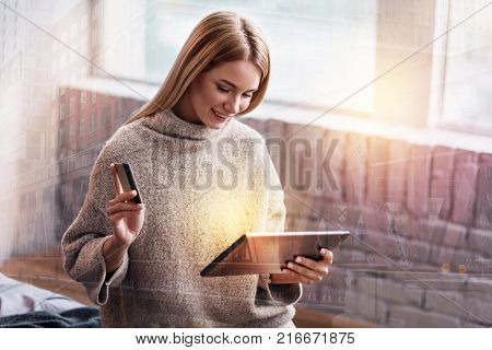 Modern gadget. Happy pleasant nice woman holding a tablet and smiling while holding a credit card
