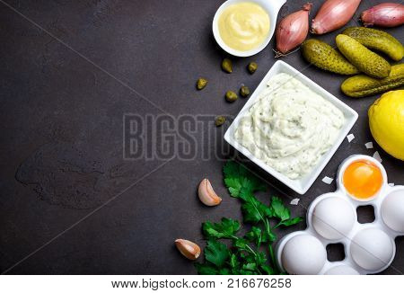 Homemade traditional french sauce remoulade in white bowl with ingredients, lemon, eggs, shallot onion, pickles, parsley, mustard on a black dark stone background. Top view, horizontal, copy space