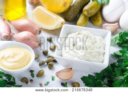 Homemade traditional french sauce remoulade in a white bowl with ingredients lemon eggs shallot onion pickles parsley mustard on a light stone background. Horizontal top view copy space