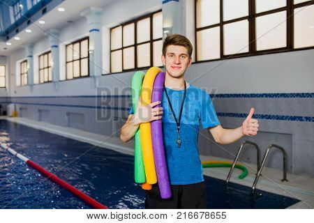 Fit smiling man holding foam roller and showing thumbs up looking at camera at swimming pool. portrait swimming instructor