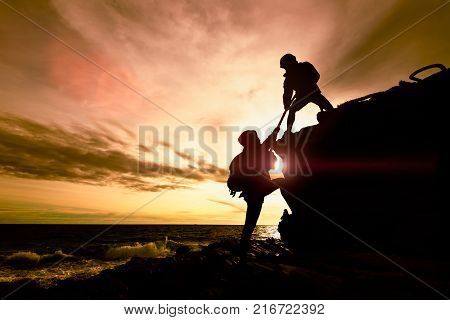 Silhouette of couple helping each other while climbing up on rocks beach in sunset. The concept of aid.