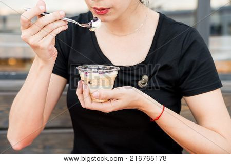 Close up female hands holding spoon and plastic glass with delicious dessert with blueberries and custard