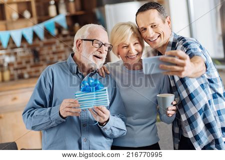 Seizing the moment. Cheerful young man taking selfies with his elderly parents during the celebration of the fathers birthday while the man holding a gift box