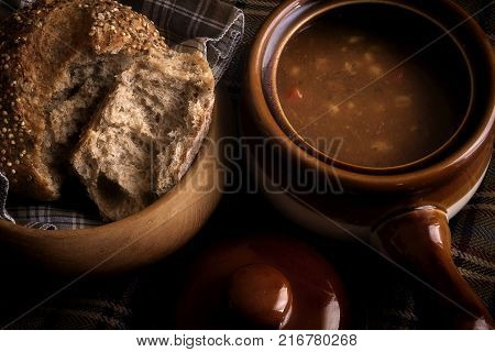 Food, a soup broth  and seeded Roll winter warming lunch, showing a rustic food scene with wooden bread dish and earthenware crockery soup dish with lid.