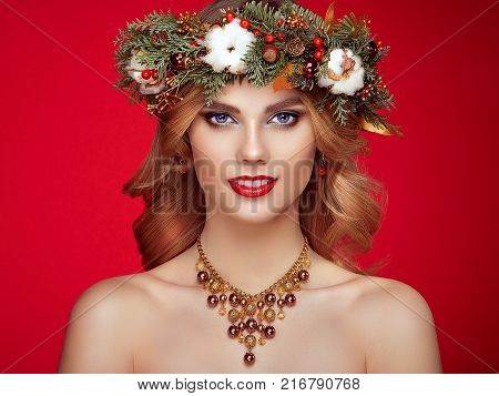 Portrait of beautiful young woman with Christmas wreath. Beautiful New Year and Christmas tree holiday hairstyle and makeup. Beauty girl portrait isolated on red background. Colorful makeup and hair