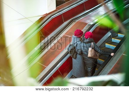 Back view two girls moving up on escalator in subway. Escalator in metro. Moving up staircase. electric escalator. View through the greenery. Selective soft focus