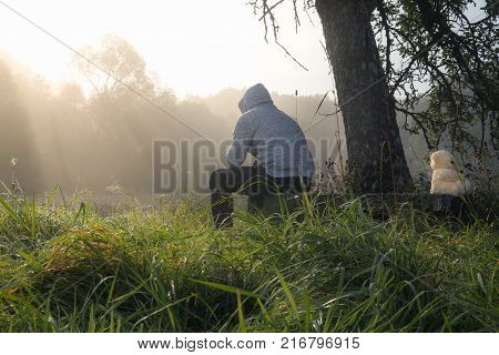 Man and teddy bear sitting on a bench - Relationship issues image with a man and a teddy bear sitting on a wooden bench under a tree while enjoying the morning mist and sun rays coming through it.