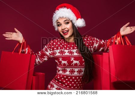 Young pretty woman or girl with long beautiful hair in Santa hat and New Year's dress holding red package on red studio background. christmas sale. Emotional brunette