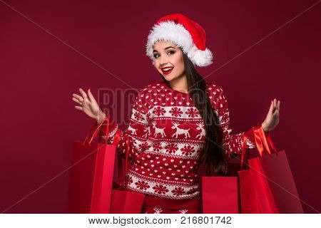 Young pretty woman or girl with long beautiful hair in Santa hat and New Year's dress holding red package on red studio background. Christmas sale. Emotional brunette. Copy space