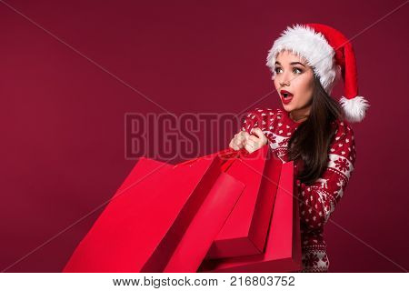 Young pretty woman or girl with long beautiful hair in Santa hat and New Year's dress holding red package on red studio background. Christmas sale. Emotional brunette. Copy space