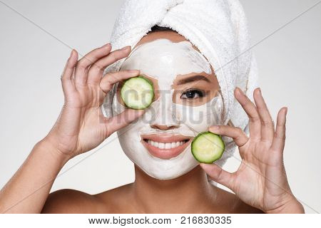 Attractive smiling asian lady with scrab on face and towel on head holding cucumber slices isolated over grey