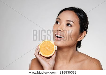 Attractive half-naked asian woman amiling while holding orange slices near her face isolated over grey