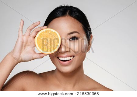 Attractive half-naked asian woman amiling while holding orange slices near her face isolated over grey