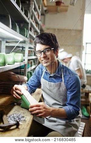 Young smiling shoe repairman sitting by workplace and changing sole of footwear