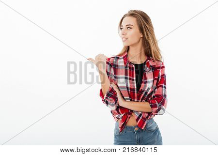 Portrait of a young casual girl in plaid shirt standing and pointing finger away at copy space isolated over white background