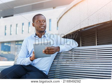 Happy businessman working on tablet. Insuranse agent browsing news on digital device, sitting on the bench outdoors near modern office center in urban area city scape