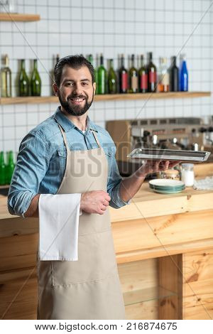 Cheerful waiter. Skilled bearded smiling waiter looking confident while being at work and being ready to bring the order with a tray in his hand