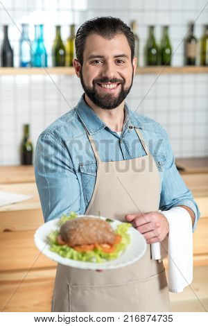 Delicious meal. Positive reliable skilled waiter looking happy while standing in a comfortable cafe and holding a white plate with a tasty meal
