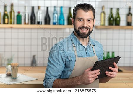 Convenient gadget. Positive emotional bearded waiter looking glad while standing with a modern convenient tablet in his hands and smiling