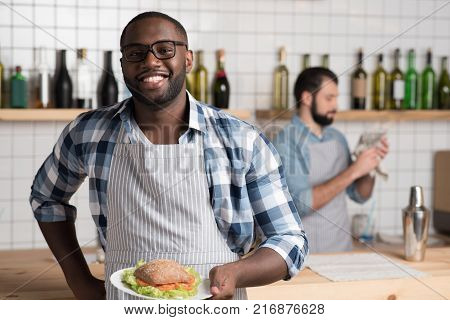 Beautiful meal. Emotional handsome experienced waiter standing at the bar counter and holding a plate with a big tasty sandwich while his colleague cleaning the classes behind his back