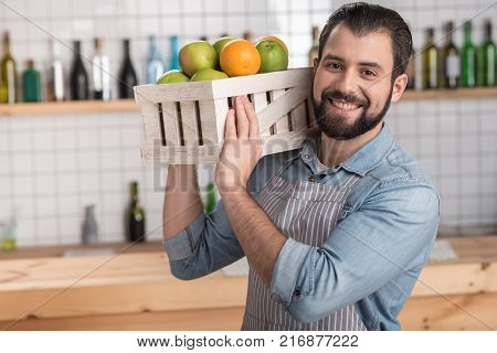 Fresh fruit. Handsome responsible enthusiastic worker looking not tired while holding a heavy wooden box with tasty fresh fruit and smiling cheerfully