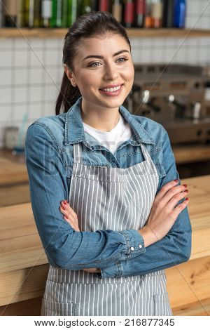 Smiling waitress. Experienced professional young waitress standing next to the bar counter with her arms crossed while having a short break before going back to the visitors of a cafe