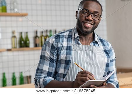 Cheerful mood. Pleasant young handsome waiter having a productive day at work while standing and making notes in a special notebook