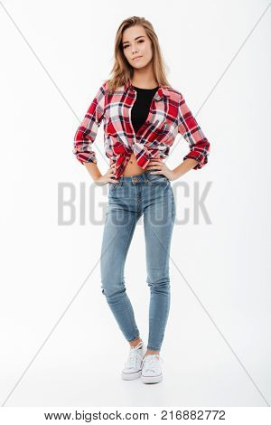 Full length portrait of a smiling casual girl in plaid shirt standing with arms on hips and looking at camera isolated over white background