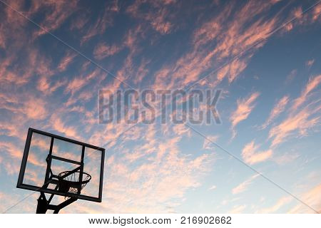 Silhouette of outdoor basketball goal with clear backboard and sunset in the background visible through backboard