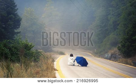 Young female in a raincoat and umbrella on the road in the fog. Travel of women in the raincoat hitchhiking in the rain
