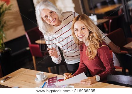 Smart women. Enthusiastic cheerful young designer looking happy while working with her older experienced creative colleague with color palettes covering their table
