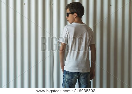 Back view. Young hipster boy dressed in white t-shirt is stands indoor against white wall. Mock up. Space for logo, text, image.