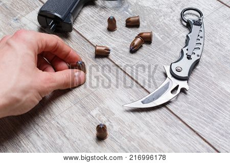 A hand holds a bullet next to other bullets, a pistol and a folding knife on a wooden floor