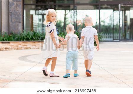 Little girl and two boys holding hands and walking outdoors with building in background. Girl is turning round and looking at camera. Back view.